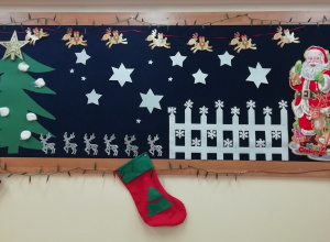 Na granatowym tle dekoracja świąteczna. Po lewej stronie znajduje się zielona choinka z żółtą gwiazdą. Po prawej stronie obok białego płotka stoi Mikołaj.
